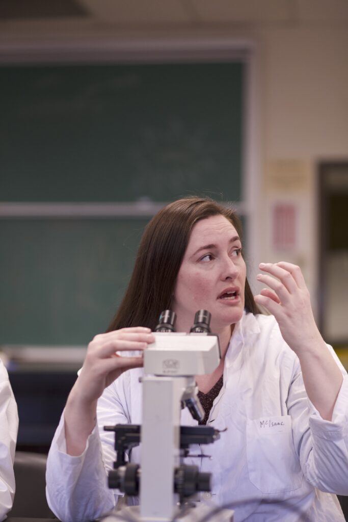 Indigenous student in a biology lab at a microscope explaining something.
